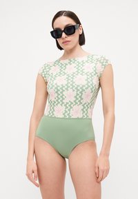 Maillot de bain une pièce avec un motif floral vert sur la partie supérieure et un bas vert uni. Comprend des manches courtes et un design à jambe haute.