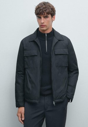 Schwarze Jacke mit Kragen, zwei Fronttaschen und einem Reißverschluss. Über einem gerippten schwarzen Pullover getragen, hat sie eine glatte Textur.
