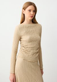 Top beige a maniche lunghe con tessuto testurizzato e stropicciato, scollatura rotonda e vestibilità slim. Progettato con un dettaglio arricciato in vita.