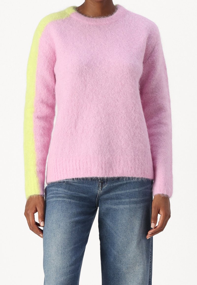 Femme portant un pull rose duveteux avec une bande jaune sur la manche gauche et un jean bleu, montrée du cou aux hanches.