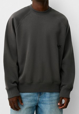 Tmavě šedá mikina s kulatým výstřihem a dlouhými rukávy, kterou nosí osoba v světle modrých džínách na jednobarevném pozadí.