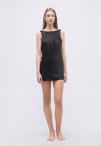 Νεαρή γυναίκα στέκεται ξυπόλητη φορώντας μαύρο μίνι φόρεμα χωρίς μανίκια μπροστά σε απλό λευκό φόντο.