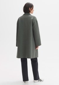 Grüner Wollmantel mit Kragen, oversized Schnitt und langen Ärmeln, kombiniert mit schwarzen Hosen und weißen Sneakers. Rückansicht vor einem weißen Hintergrund.