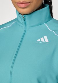 Tyrkysová športová bunda s vysokým golierom, zipsom, bielym kontrastným lemovaním a potlačeným logom. Tkanina sa zdá byť hladká a elastická.