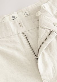 Pantalon beige clair avec fermeture éclair et bouton, en tissu texturé. Comprend une étiquette de marque à l'intérieur de la ceinture.
