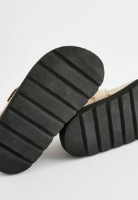 Chaussures beiges avec une semelle en caoutchouc noir texturé, dotée de rainures profondes pour une meilleure traction. La partie supérieure est en tissu doux avec une finition neutre.