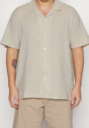 Mand iført en lysebeige kortærmet skjorte med knapper og matchende beige shorts, med en blomster-tatovering på venstre underarm.