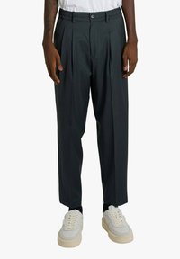 Uomo che indossa pantaloni pieghettati grigio scuro, sneaker bianche e una camicia bianca, in piedi con le braccia rilassate ai lati.