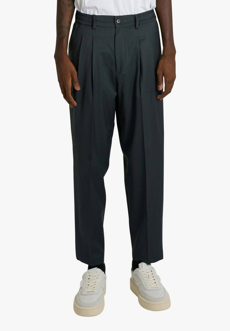 Uomo che indossa pantaloni pieghettati grigio scuro, sneaker bianche e una camicia bianca, in piedi con le braccia rilassate ai lati.