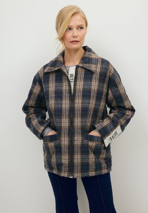 Vrouw draagt een blauw-beige geruit jack met rits en kraag, handen in de zakken, en een donkere broek tegen een effen lichte achtergrond.