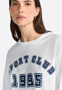 Biela bavlnená tričko s potlačou "SPORT CLUB 1995" v námorníckej modrej, okrúhly výstrih a voľný strih. Hladká textúra a neformálny dizajn.