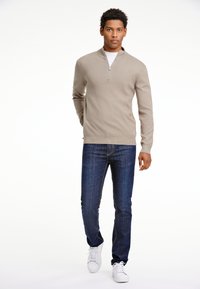 Beige zip-up neule, jossa on ribbed hihansuut ja helma, yhdistetty tummansinisiin farkkuihin ja valkoisiin tennareihin, seisoo valkoista taustaa vasten.