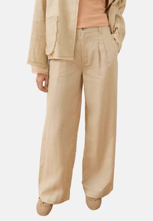 Personne portant un pantalon beige à jambes larges, une veste assortie, un haut couleur pêche et des chaussures beiges sur un fond uni.