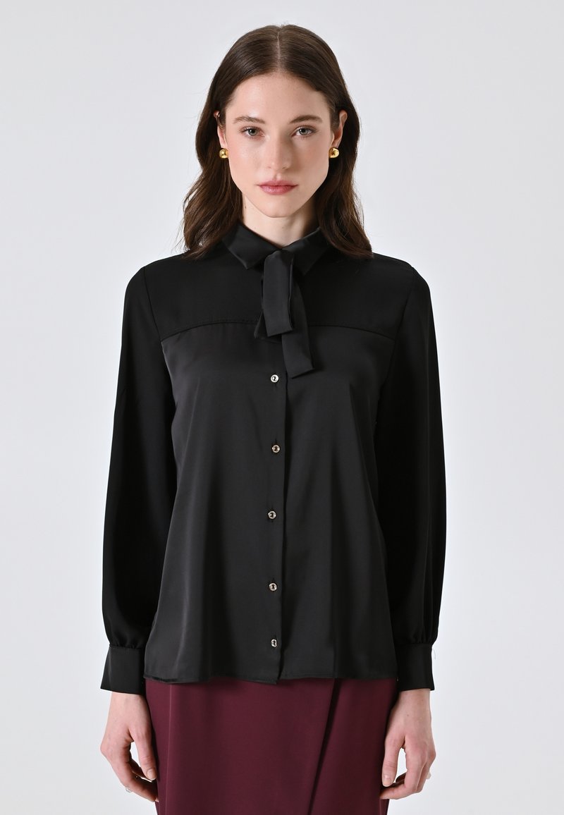 Schwarze Bluse mit Kragen und gebundener Schleife, mit langen Ärmeln und Knopfverschluss. Glatte Textur mit glänzenden Akzenten, mehrlagiges Design.