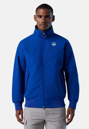 Giubbotto bomber blu con collo alto, chiusura a zip e tasche laterali. Presenta un logo circolare sul petto e polsini e orlo a coste.