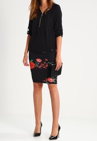 Black blouse with zipper detail and rolled sleeves, paired with a fitted pencil skirt featuring floral patterns in red and green. Black clutch and heels.