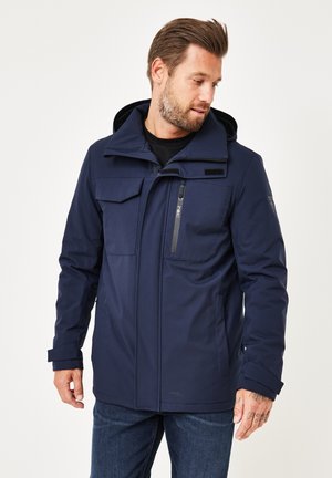 Man wearing navy blue hooded jacket with multiple pockets and jeans, looking down to the side against a plain white background.