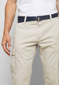 Pantalones cargo beiges con bolsillos laterales, llevados con un cinturón trenzado azul marino con hebilla metálica. La tela parece ligera y texturizada.