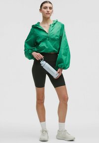 Green lightweight hooded jacket, black shorts, white shoes with matching ankle socks, holding a light blue water bottle with logo.