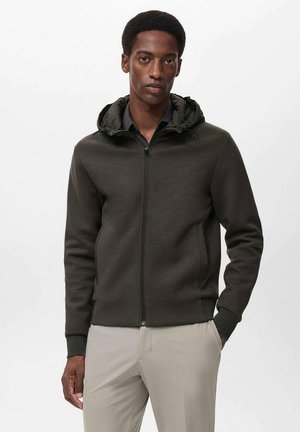 Donkergroene rits hoodie met een gestructureerde afwerking, voorzien van een capuchon en voorzakken, gedragen over een zwart shirt en lichtgrijze broek.