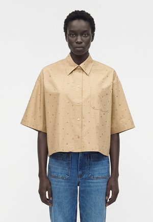 Άτομο που φορεί ένα μπεζ πουκάμισο με κοντά μανίκια με μικρές διακοσμήσεις και μπλε τζιν με μεγάλες μπροστινές τσέπες, στέκεται αντικριστά.