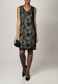 Černé krajkové šaty bez rukávů s výstřihem do V, zdobené květinovými vzory a podélným lemem. Odpovídají s malou černou kabelkou.