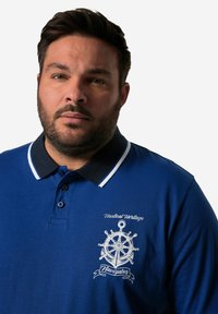 Modrá polo košile s námořnickým límcem a bílými akcenty. Obsahuje bílé vyšívané námořnické logo a text na levé straně.