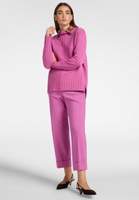 Maglione rosa lavorato a maglia con collo alto e bordo a coste, abbinato a pantaloni rosa coordinati e mule con tacco marroni. Texture liscia.