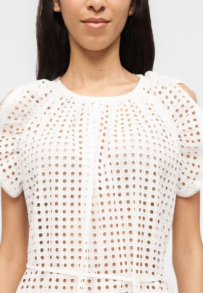 Γυναίκα που φοράει λευκό φόρεμα με κοντά μανίκια, σχέδιο δαντέλας με τετράγωνη φοδραριστή κοπή και λεπτή ζώνη στη μέση, εμφανιζόμενη από τους ώμους μέχρι τη μέση.