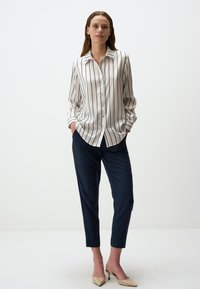 Weiß gestreiftes Hemd mit Knöpfen und Kragen, kombiniert mit marineblauen, schmal zulaufenden Hosen und hellbraunen High Heels, an einem Modell vor einem schlichten Hintergrund.