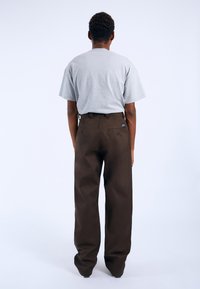 Braune weit geschnittene Hosen mit einer geknöpften Gesäßtasche, kombiniert mit einem hellgrauen T-Shirt mit kurzen Ärmeln, von hinten vor einem schlichten Hintergrund betrachtet.