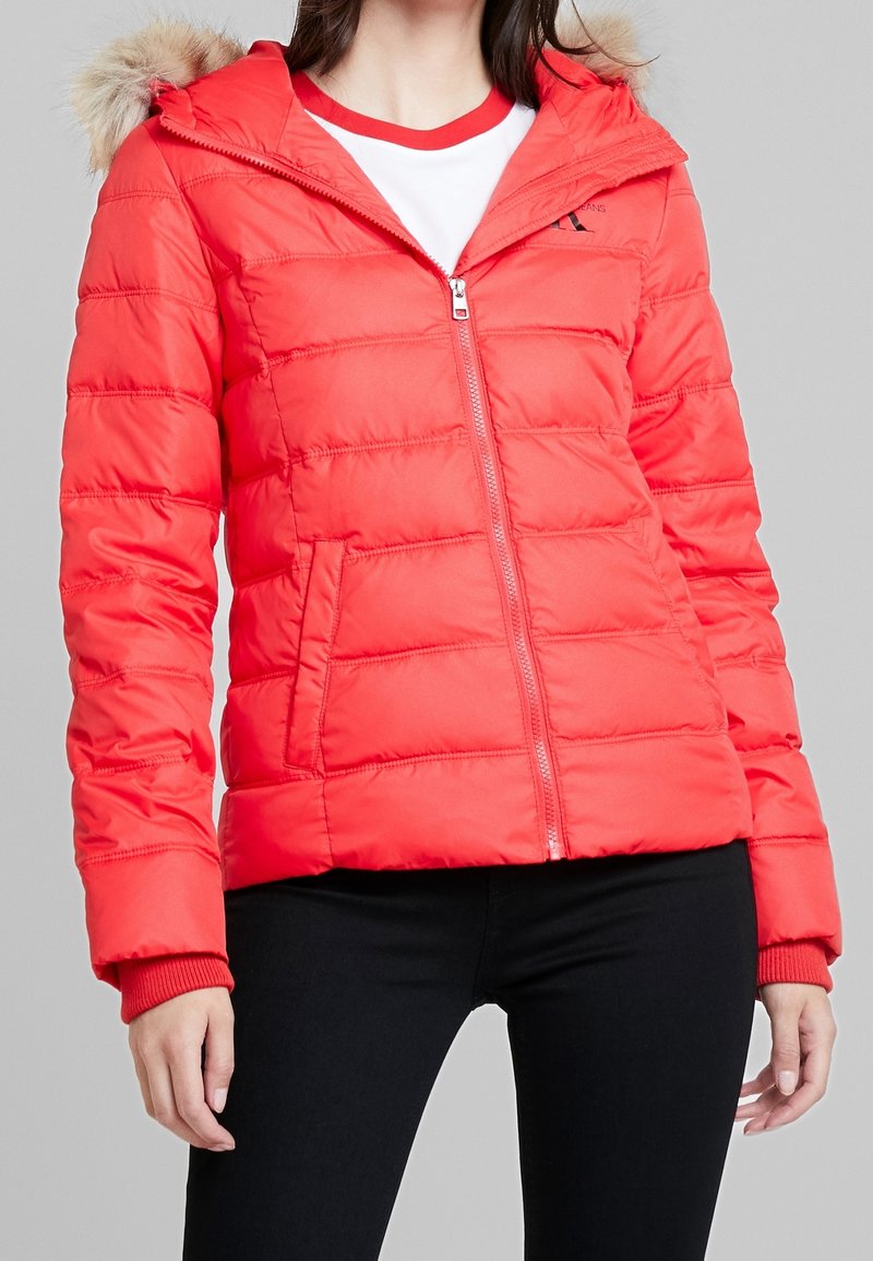Rode donzenjas met een capuchon gevoerd met bont, ritssluiting en horizontale stiksels. Voorzien van zijzakken en contrasterende gebreide manchetten.