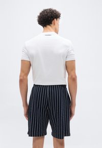 Άνδρας με κοντά σγουρά μαλλιά, που φοράει λευκό t-shirt και μαύρη βερμούδα με κάθετες λευκές λωρίδες, στέκεται με την πλάτη προς το μέρος μας σε λευκό φόντο.