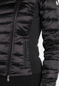 Gros plan sur une veste matelassée noire avec des panneaux en tricot sur les côtés, fermeture éclair à l'avant et poche zippée sur le côté gauche.