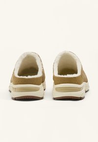 Brune ruskind slip-on sko med en fluffy hvid foer, der har en tyk cremefarvet mellemsål og en gummiydersål. Bagfra vises den polstrede hæl.