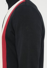 Zwarte trui met witte en rode diagonale strepen, met een gestructureerde stof en een hartvormig logo geborduurd op de mouw.