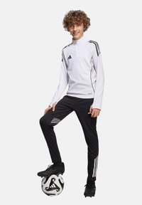 Haut de sport zippé blanc avec des rayures noires, associé à un pantalon noir avec des accents gris. Des crampons de foot noirs et un ballon sont inclus.