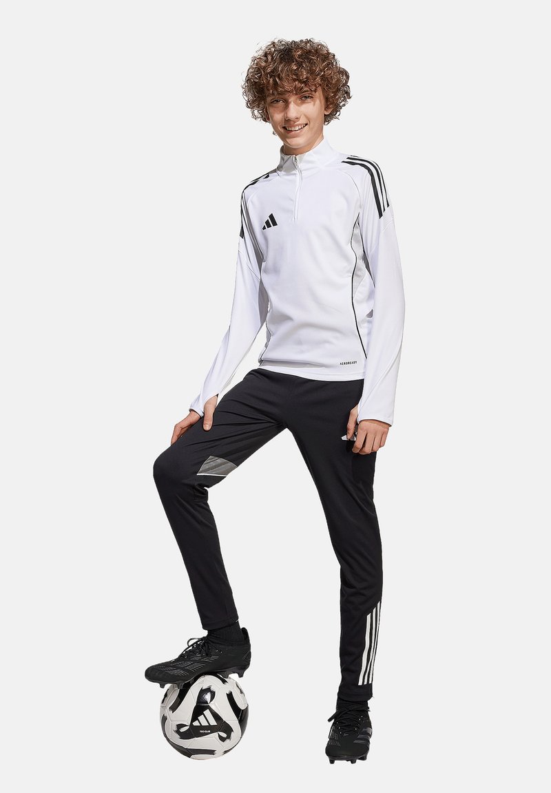 Haut de sport zippé blanc avec des rayures noires, associé à un pantalon noir avec des accents gris. Des crampons de foot noirs et un ballon sont inclus.