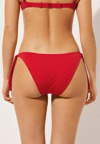 Červený bikinyspodní díl s vázacím designem po stranách. Hladká tkanina s minimálním pokrytím, s kulatými okraji a štíhlou siluetou.