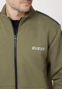Giacca a zip verde oliva realizzata in tessuto morbido, con colletto alto, accenti a strisce nere sulle spalle e un logo "GUESS" bianco.