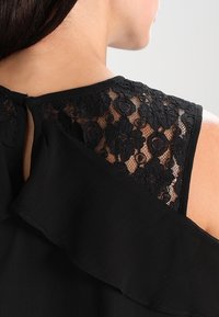 Top nero con un design arricciato e dettagli in pizzo sulle spalle. Il tessuto è strutturato, creando un contrasto visivo con il pizzo.