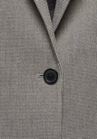 Blazer gris con un fino patrón de cuadros, cierre de botón negro y una textura suave con un corte moderno.