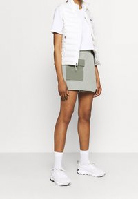 Chaleco acolchado blanco sobre una camiseta blanca, combinado con pantalones cortos verde olivo con un gran bolsillo frontal, y zapatillas deportivas blancas con detalles en gris.