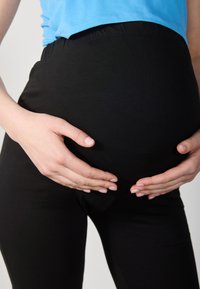Juodi tamprūs nėštumo leggingai su aukštu juosmeniu. Žmogaus rankos ilsisi ant apvalaus pilvo, dėvintis šviesiai mėlyną marškinėlį. Sklandi tekstūra.