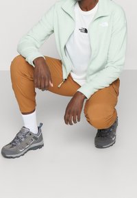 Giacca a zip verde chiaro, t-shirt bianca con logo nero, pantaloni marroni, scarpe da trekking grigie con accenti blu, in posizione inginocchiata su uno sfondo neutro.