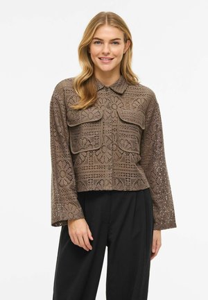 Femme souriante portant une chemise marron en crochet à boutons avec deux grandes poches devant et un pantalon noir, sur un fond blanc uni.