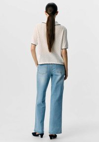 Vit plagg med öppen stickning och svarta detaljer, kombinerad med ljusblå vida jeans. Modellen har långt hår i en hästsvans. Svarta klackskor.