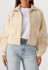 Beige wollen jasje met witte spiraalvormige borduursels, cropped ontwerp, knoopsluiting en brede mouwen, gedragen over een wit shirt en lichte jeans.
