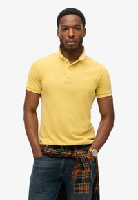Polo giallo con colletto, maniche corte e un marchio discreto. Indossato con jeans scuri e una camicia a quadri annodata in vita.