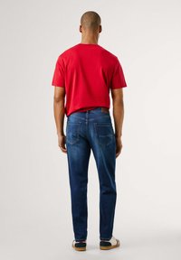 Rdeča bombažna majica s kratkimi rokavi in temno modri trakovni kavbojke, ki imajo ravno prileganje in vidne šive. Čevlji z belimi in mornarsko modrimi poudarki.